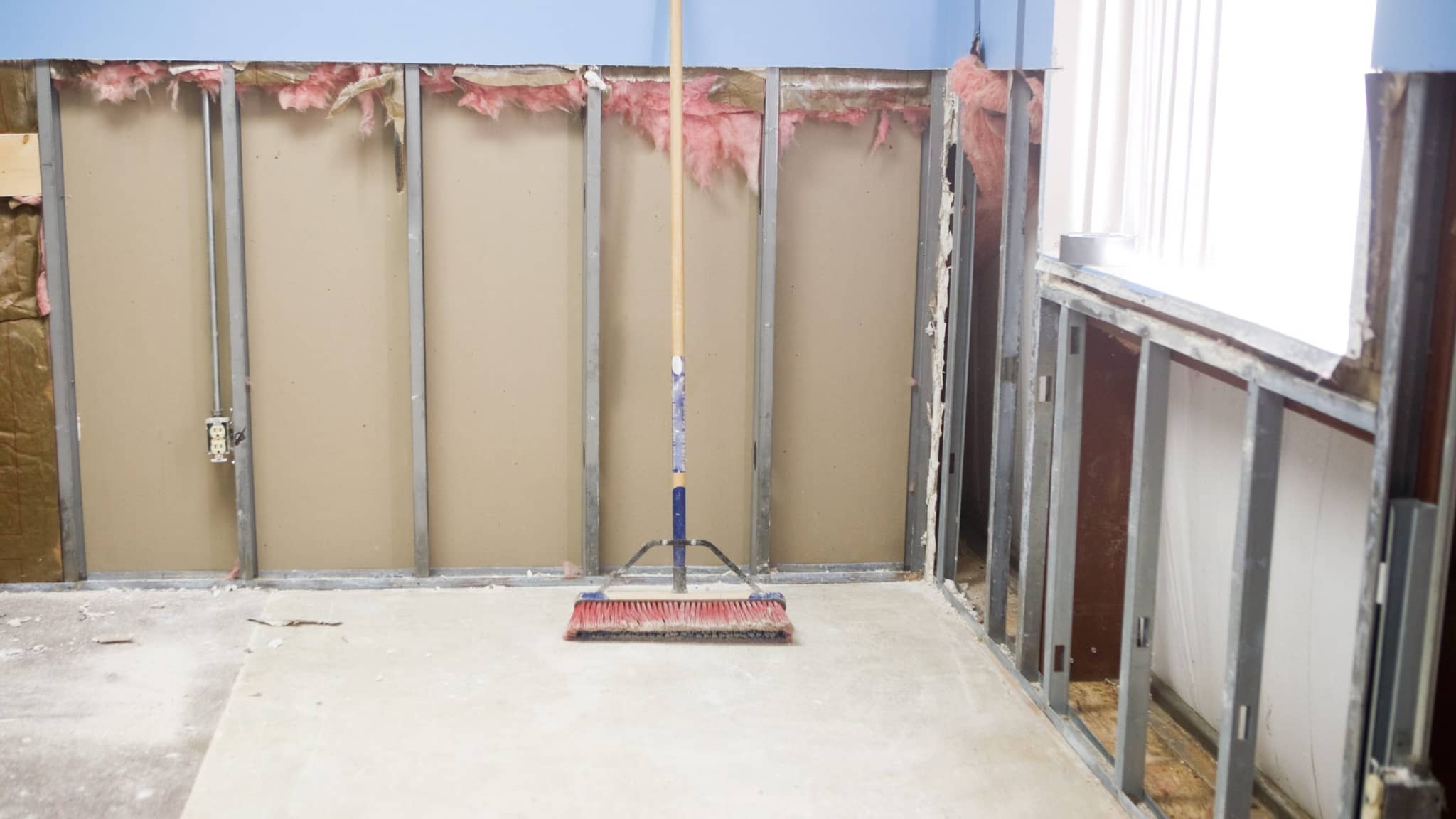 Wall cut open along the bottom with old pink insulation exposed above and a push broom on the floor