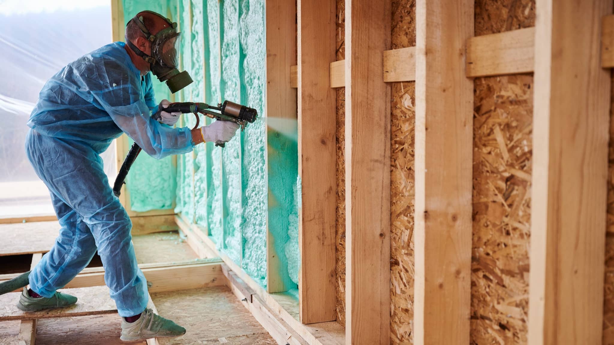 Installer in a protective suit and respirator spraying foam insulation into open wall stud bays